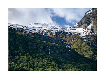 Fotografie der schneebedeckten Gipfel des Lysefjords in Norwegen mit grünen Wäldern im Vordergrund.
