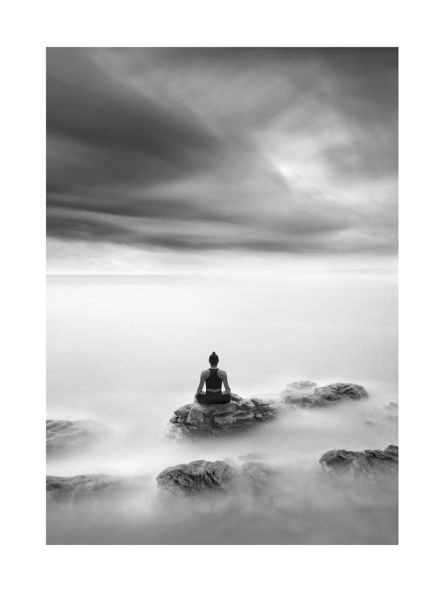 Schwarzweiß-Fotografie einer Frau, die auf einem Felsen im Meer meditiert.