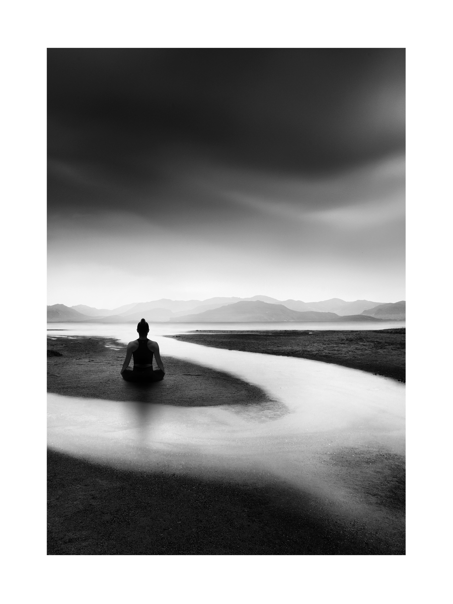 Schwarz-Weiß-Fotografie einer Frau, die am Fluss meditiert, mit Bergen im Hintergrund.