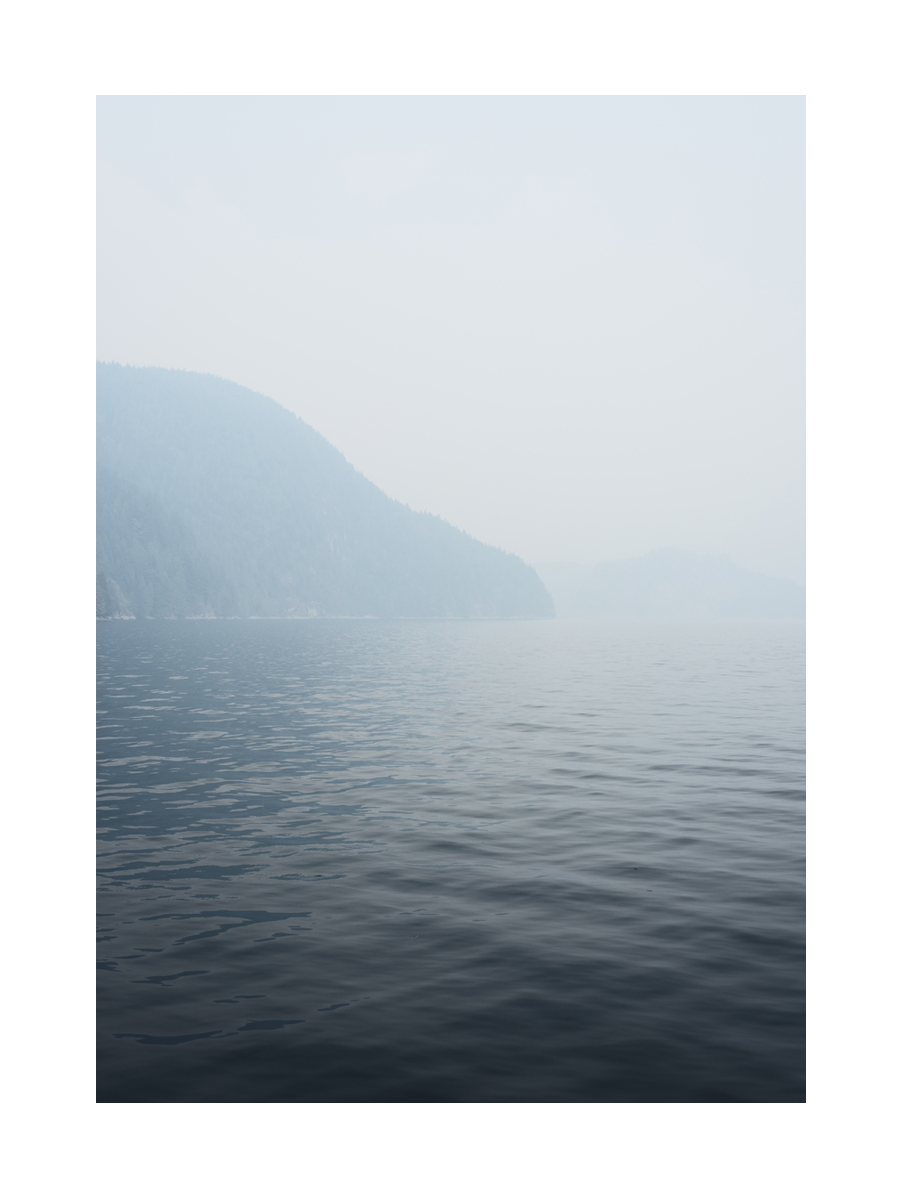 Fotografie eines nebligen Sees mit ruhiger Wasseroberfläche und bewaldeten Hügeln im Hintergrund.