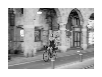 Schwarz-Weiß-Fotografie einer Frau auf einem Fahrrad in einer urbanen Umgebung mit Bögen.