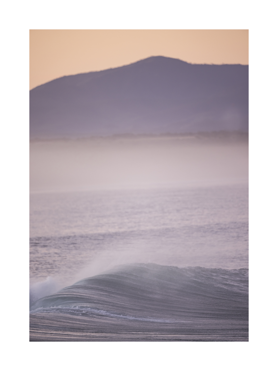 Fotografie einer Welle im Meer mit einem Berg im Hintergrund und einem rosa Himmel.