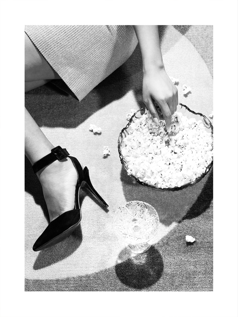 Schwarz-weiß-Fotografie eines Beins in einem Absatzschuh neben einer Schüssel Popcorn auf einem Teppich.