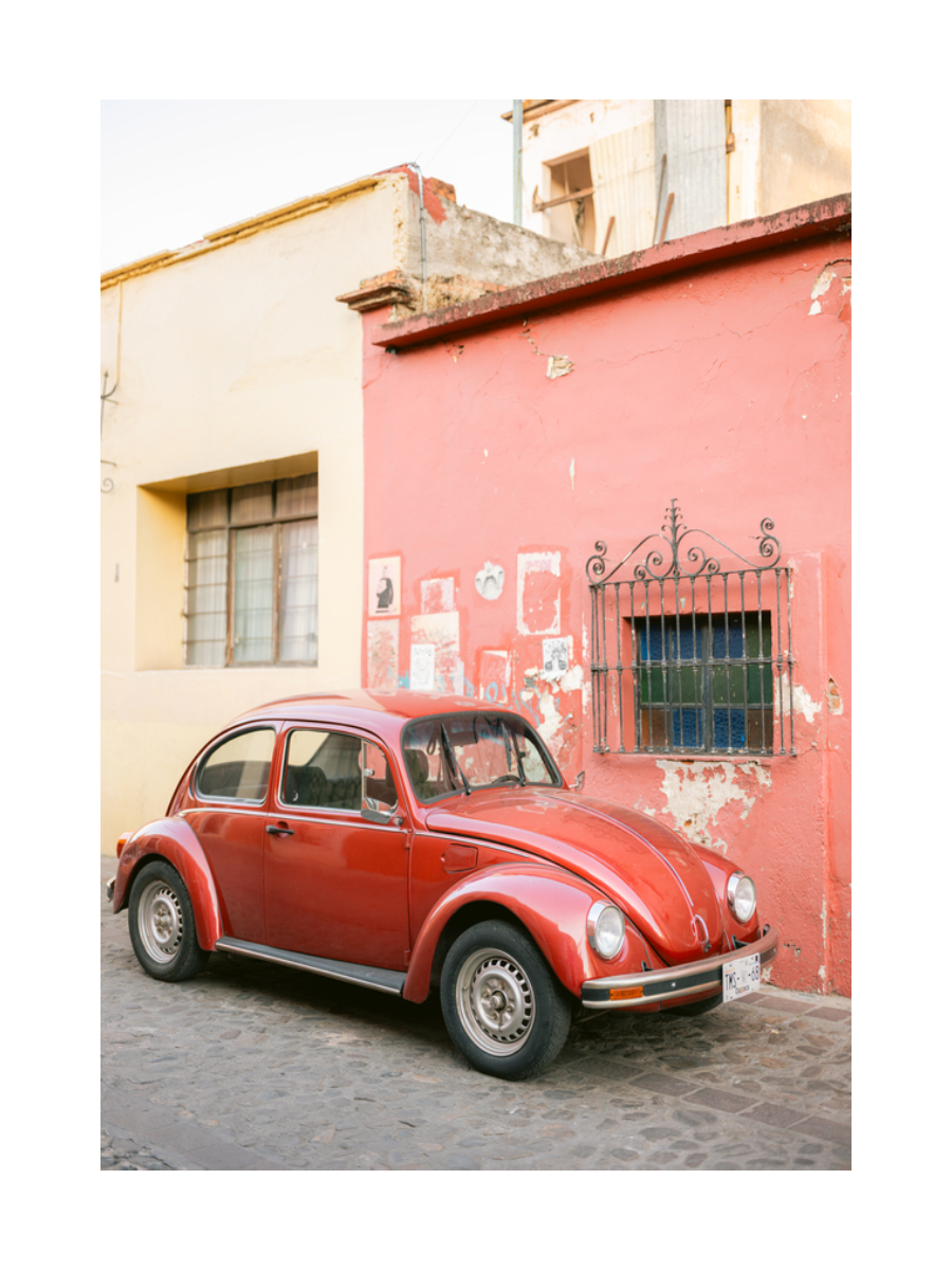 Ein roter VW Käfer neben einem Gebäude mit rosa Wand und gelbem Fenster.