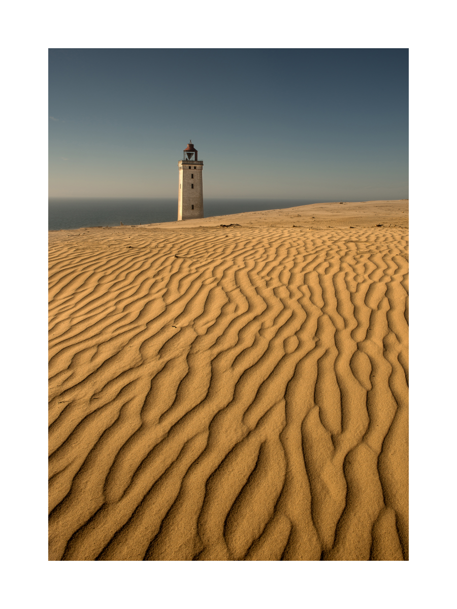 Ein Leuchtturm in einer Wüstenlandschaft mit Sanddünen und blauem Himmel.