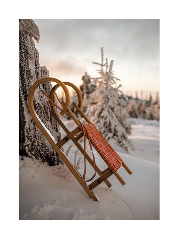 Ein Schlitten lehnt im Schnee an einem Baum, mit einem Sonnenuntergang im Hintergrund.