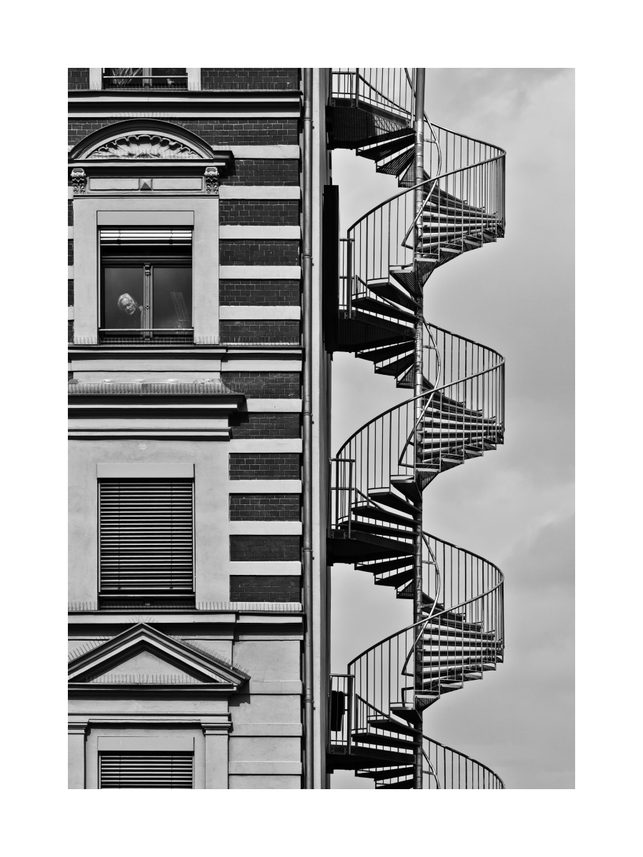 Schwarz-Weiß-Fotografie eines Gebäudes mit Wendeltreppe und einer Person im Fenster.