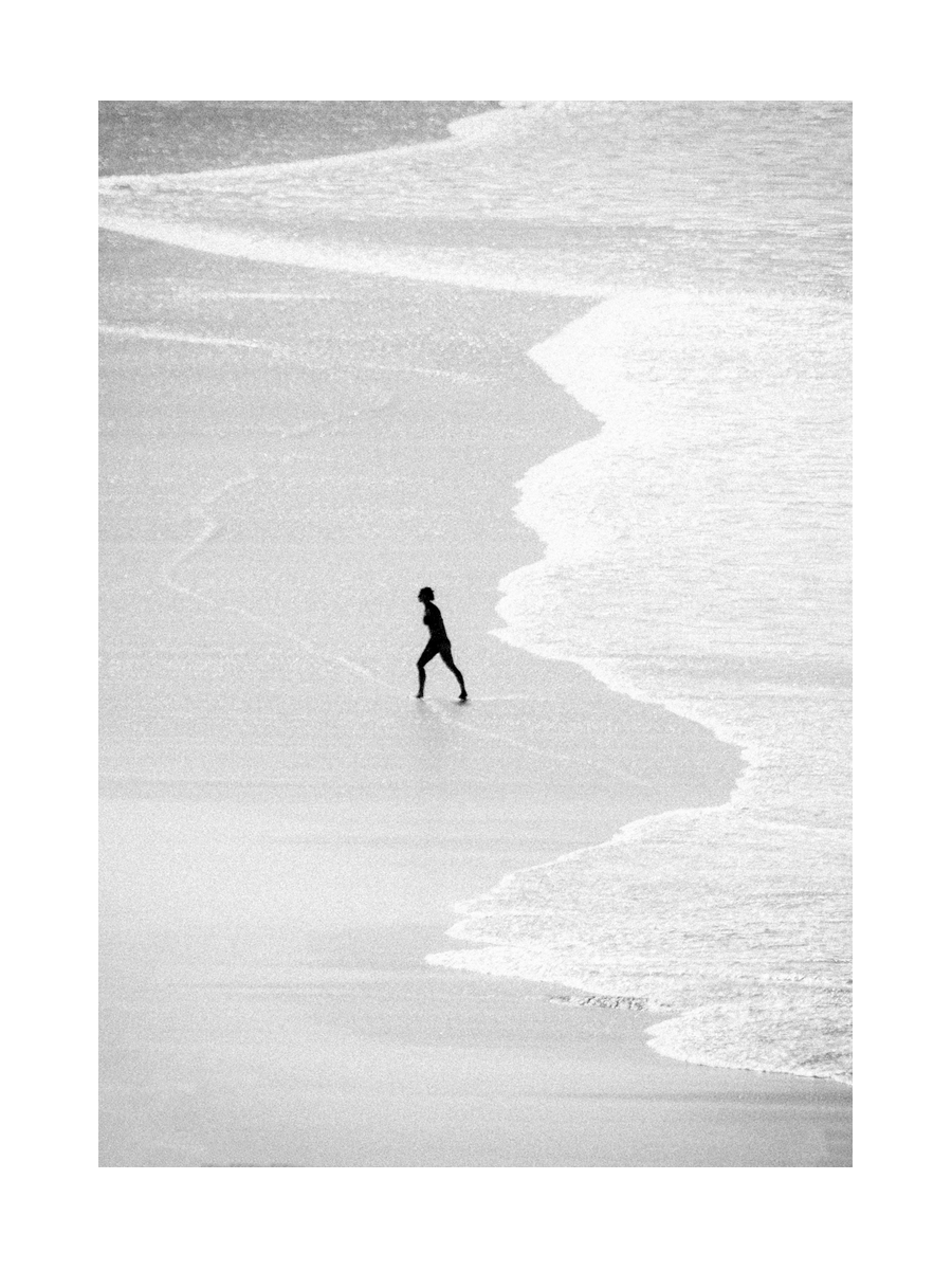 Schwarz-weiß-Fotografie eines Surfers, der am Strand entlanggeht.