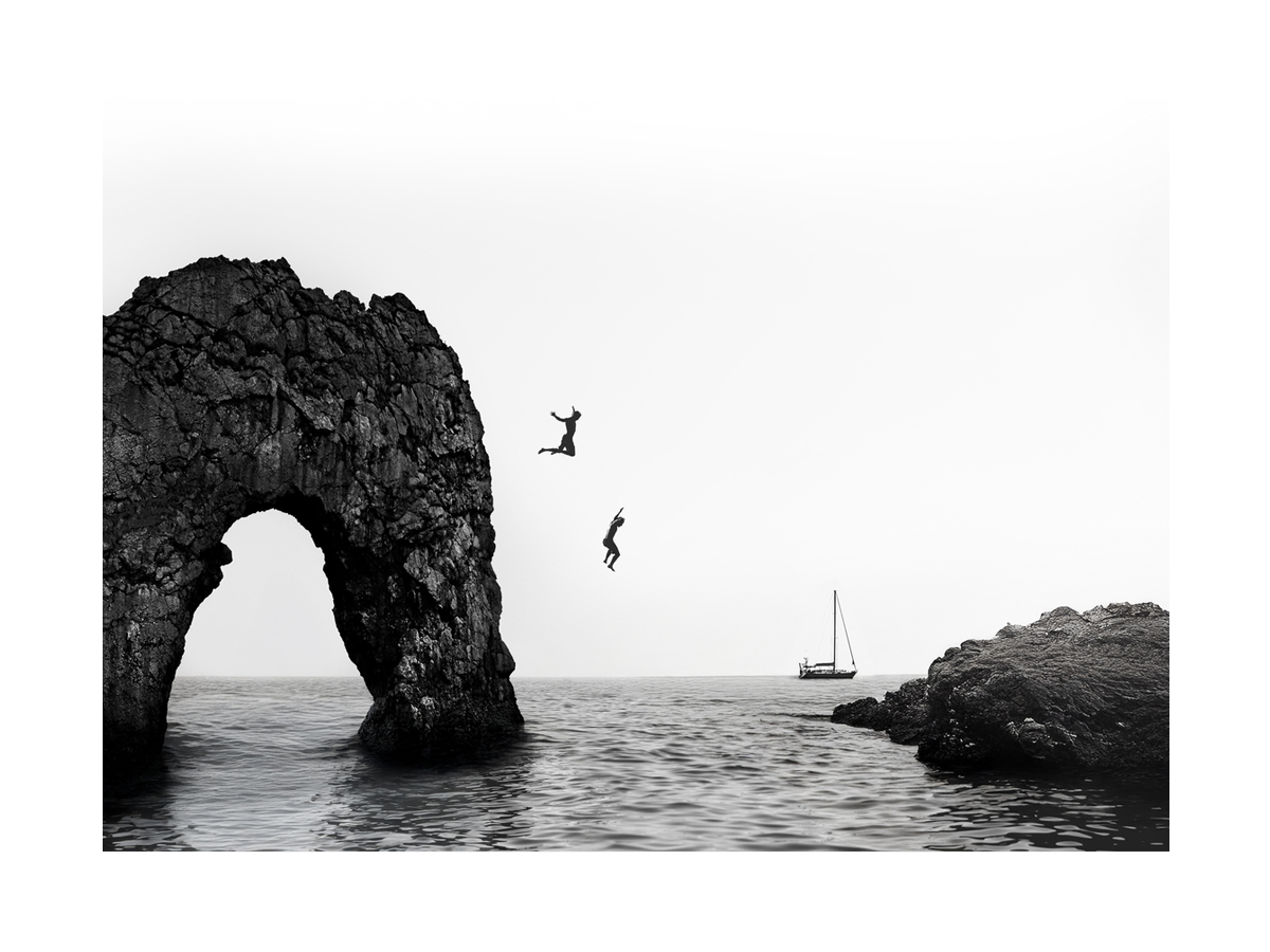 Schwarz-weißer Fotodruck von zwei Personen, die von einer Klippe ins Meer springen, mit einem Boot im Hintergrund.