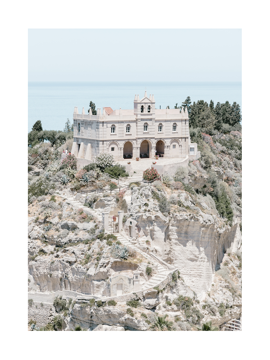 Fotografie eines beigen Gebäudes auf einem Felsen mit Treppe und Meer im Hintergrund.
