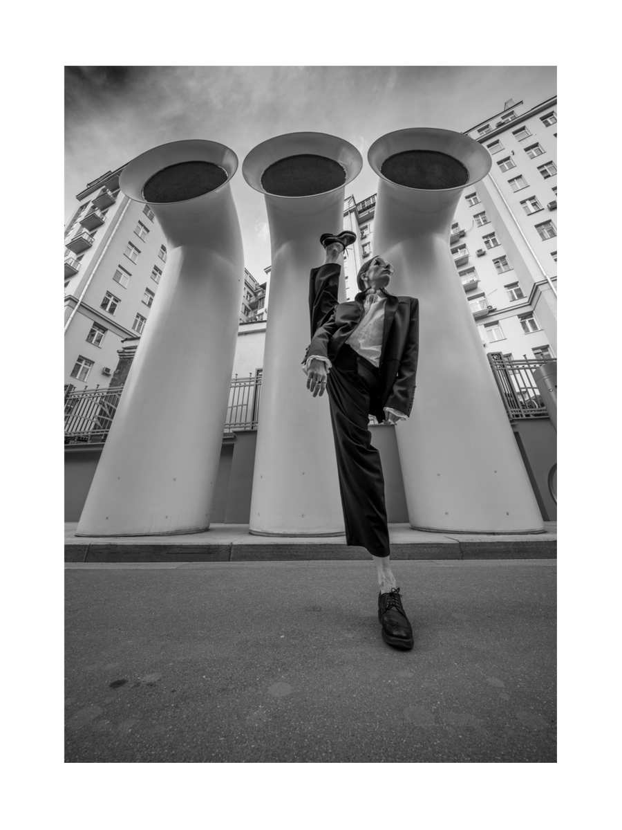 Schwarz-Weiß-Fotografie einer Person in Tanzpose vor urbaner Kulisse mit hohen, zylindrischen Strukturen.