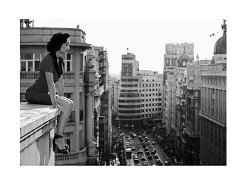 Schwarz-weiße Fotografie einer Frau in einem gepunkteten Kleid, die auf einem Dach über einer Stadt sitzt.