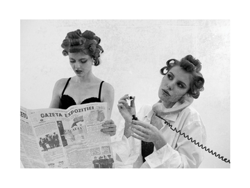 Schwarz-weiß-Fotografie von zwei Frauen mit Lockenwicklern, eine liest eine Zeitung, die andere telefoniert.