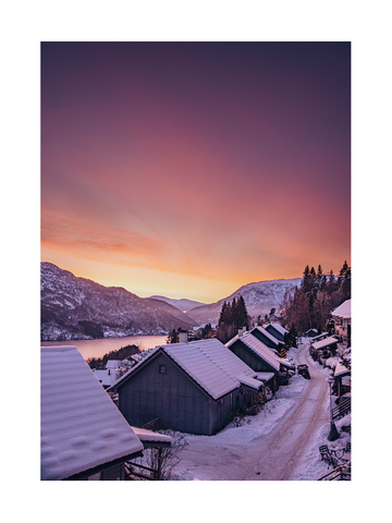 Verschneite Winterlandschaft mit Häusern und einem Sonnenuntergangshimmel in Orange- und Violetttönen.