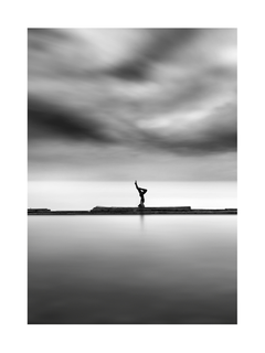Schwarz-weiß-Fotografie einer Frau, die Yoga auf einem Steg im Meer macht, mit bewölktem Himmel.