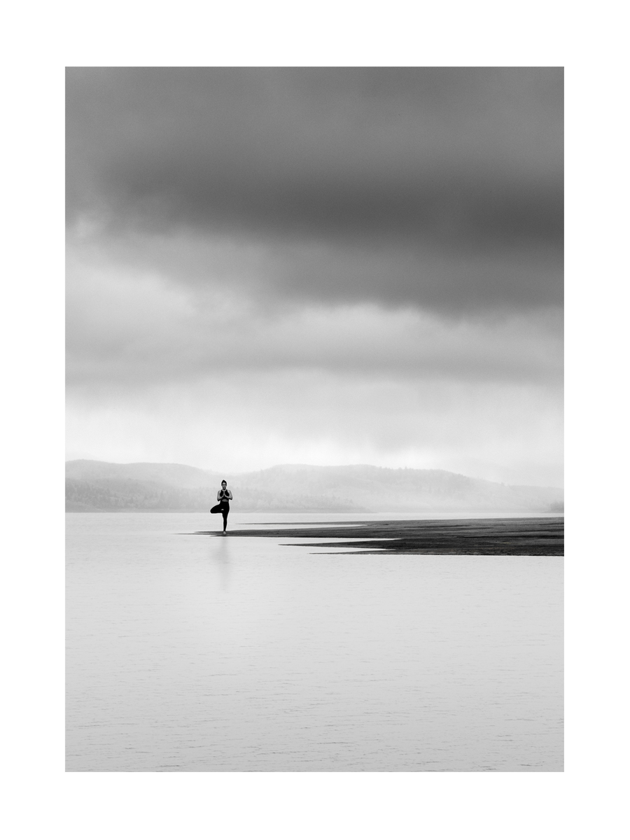 Schwarz-weiße Fotografie einer Person, die Yoga auf einem See macht, umgeben von Wasser und Wolken.
