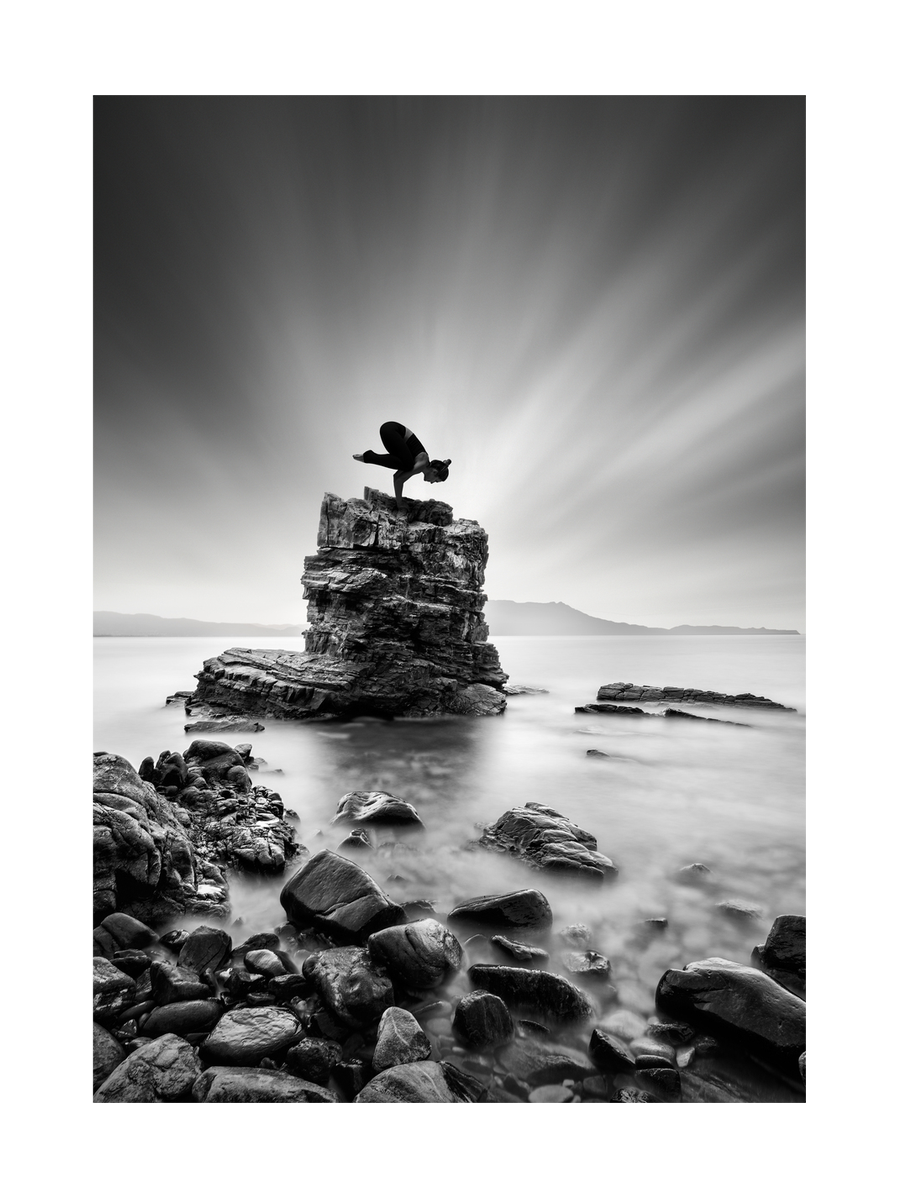 Schwarz-weiß-Fotografie einer Frau in einer Yoga-Pose auf einem Felsen im Meer.