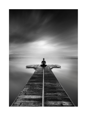 Schwarz-weiß-Fotografie einer Frau, die auf einem Steg in einem See meditiert.