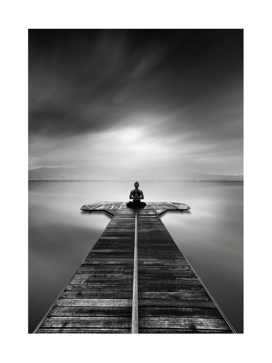 Schwarz-weiß-Fotografie einer Frau, die auf einem Steg in einem See meditiert.